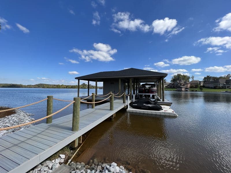 Covered boat dock with a dark roof at the end of a rope-railed walkway on a calm lake