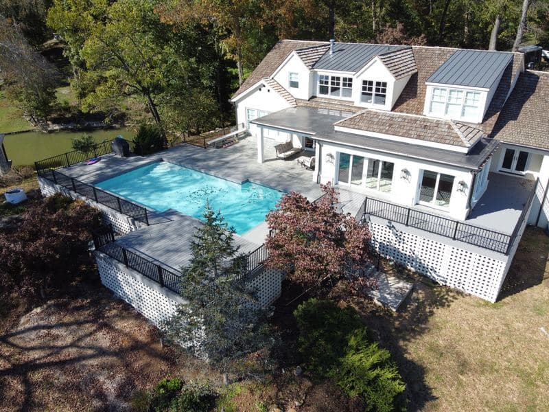 Aerial view of a gray composite pool deck wrapping a white lakeside home, with black metal railings and white lattice skirting