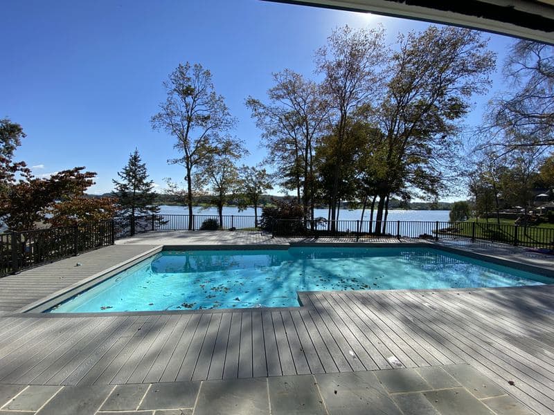 Composite pool deck with black railings overlooking the lake, framed by a stone patio and autumn trees