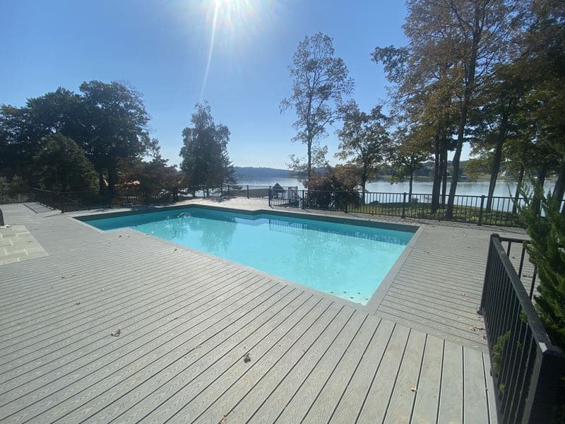 Sunlit composite deck surrounding an in-ground pool, with black metal railings and a lake view beyond