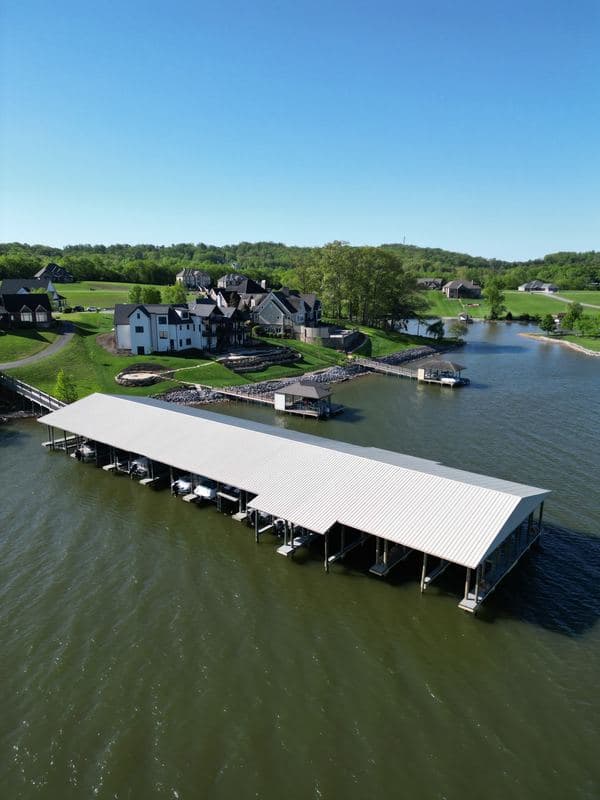 Aerial view of a long covered marina with a white metal roof and boat slips, lakefront homes beyond