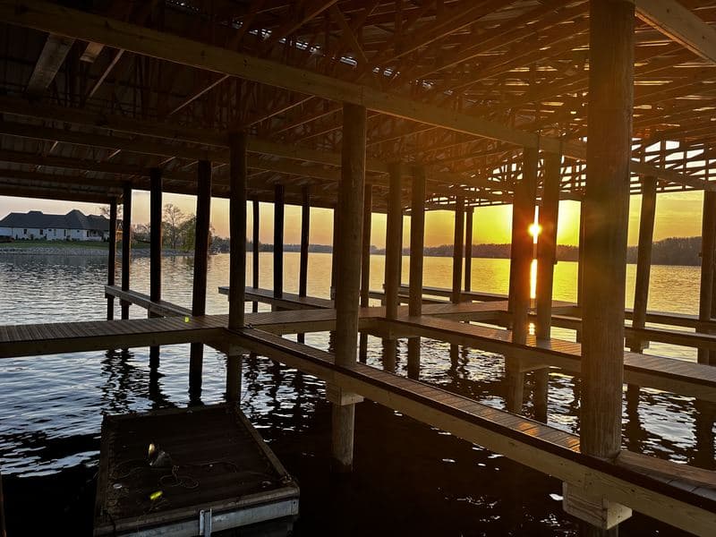 Sunset over the lake seen from beneath the marina roof, with heavy timber framing and boat slips