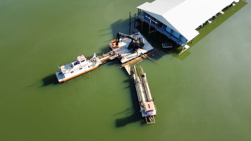 Top-down aerial of a DOCK&DECK barge, excavator, and work boat with pilings staged beside the marina