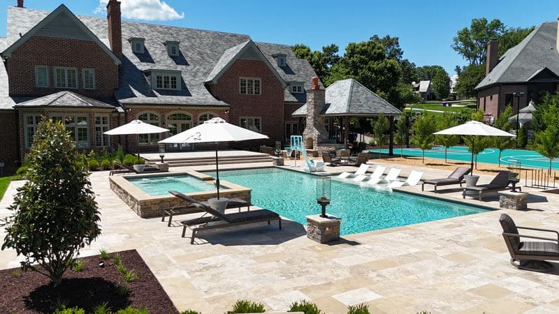 Wide view of the pool, spa, and travertine patio with umbrellas and loungers, sport court and pavilion beyond