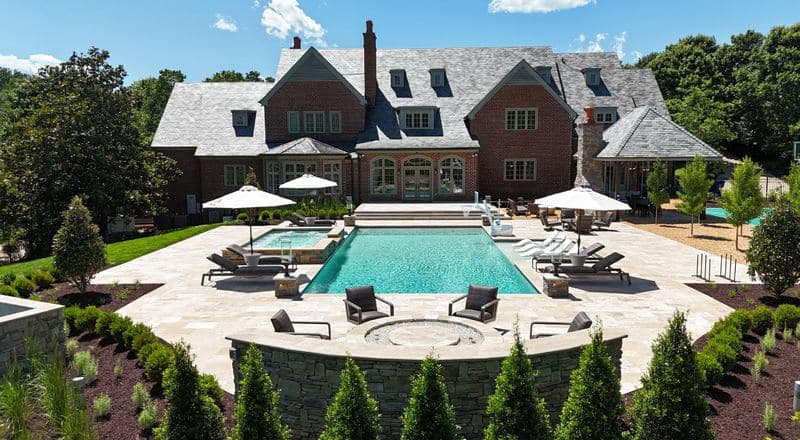 Rectangular pool and spa on a travertine patio with umbrellas and loungers, framed by a curved stone fire-pit wall