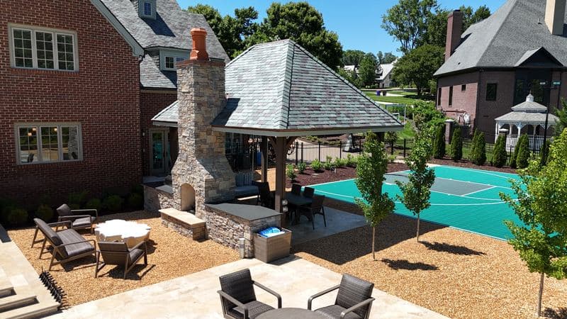 Stone-roofed pavilion with a tall stacked-stone fireplace and outdoor dining beside the teal sport court