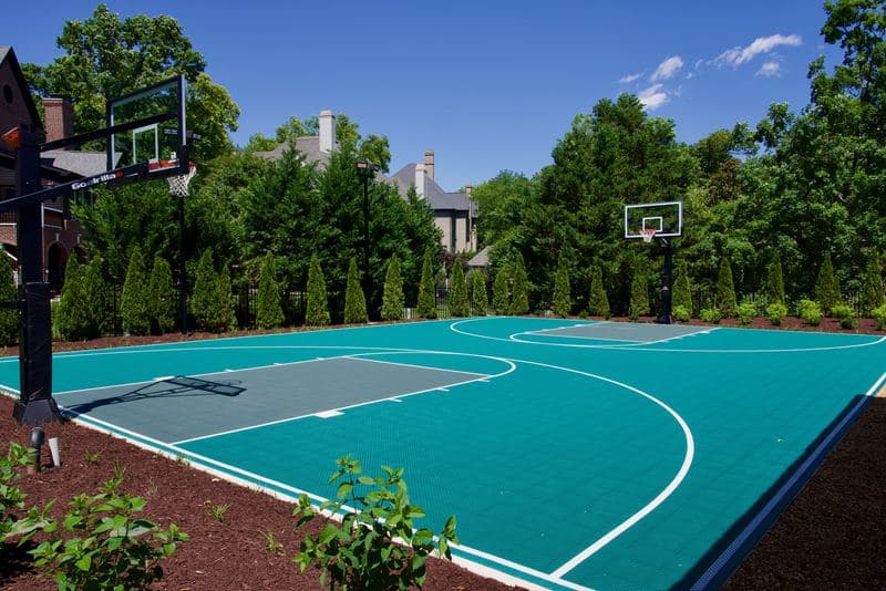Teal backyard basketball sport court with painted lines and hoops, edged by mulch beds and young shrubs