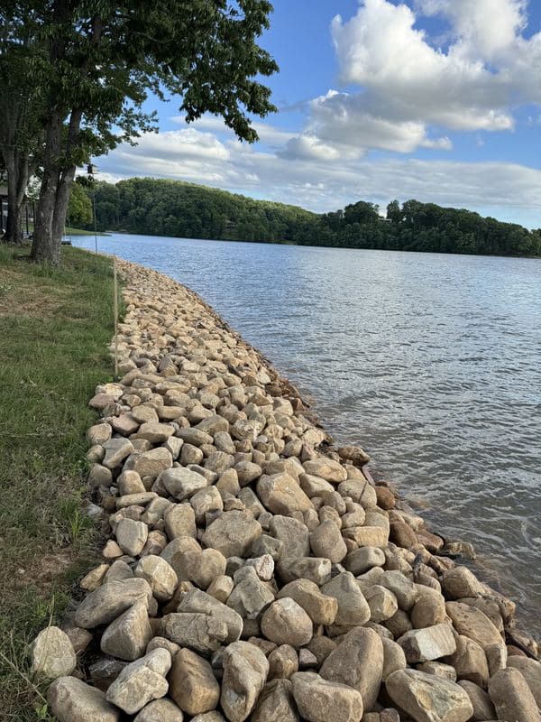 Long sweep of new river rock lining the lake shoreline beside a mowed lawn, wooded hills across the water