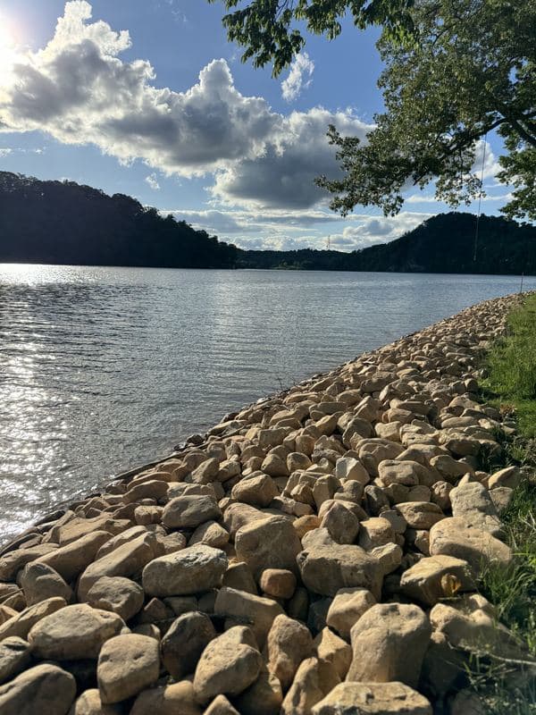 Close view of sunlit river rock armoring the lake shoreline, with hills and clouds on the horizon