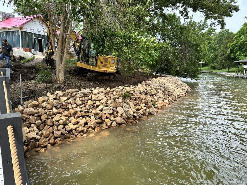 Excavator placing river rock along the lake shoreline during shoreline stabilization work