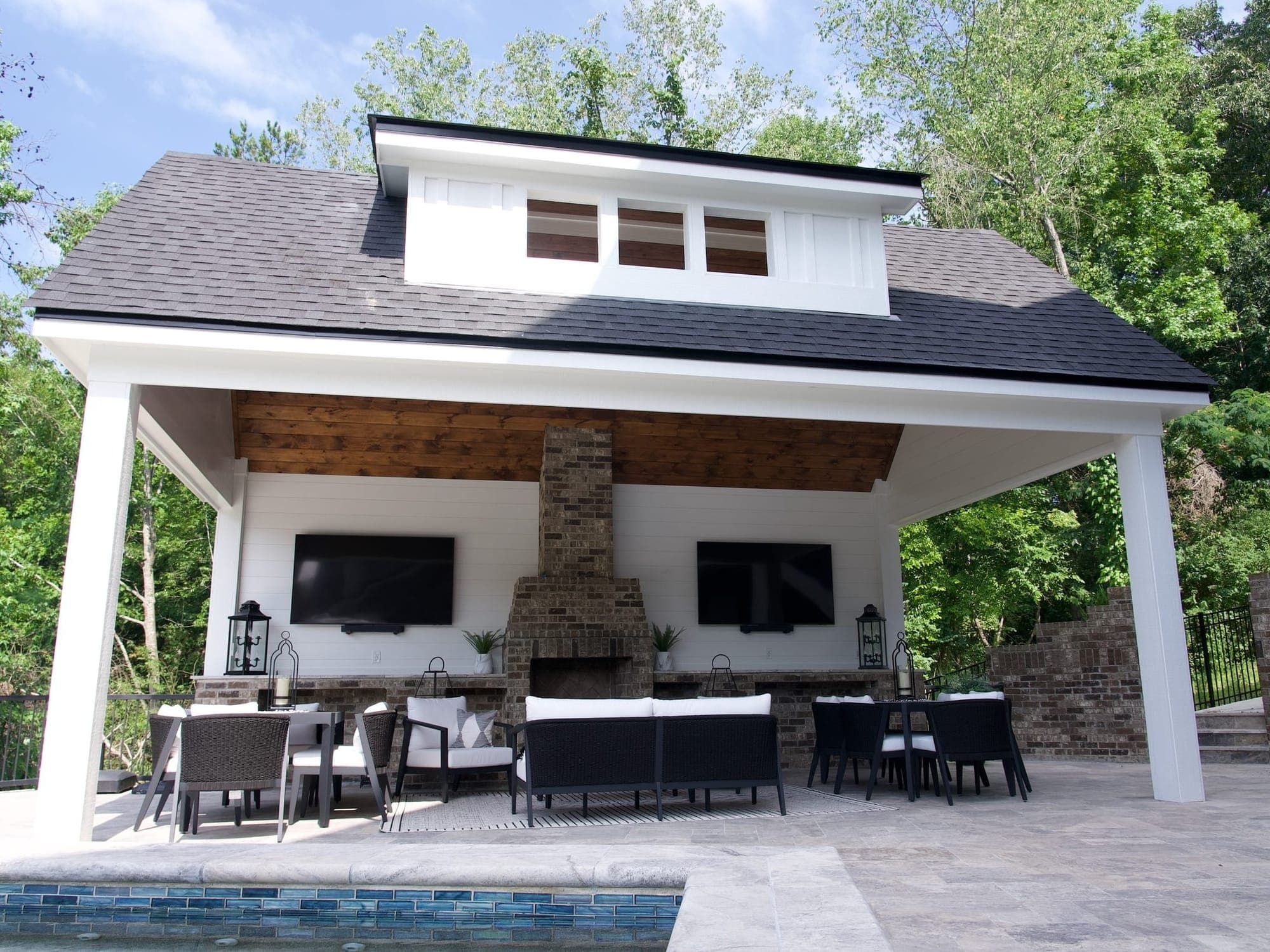 Covered poolside pavilion with a stone fireplace flanked by two wall-mounted TVs, lounge seating, and dining tables