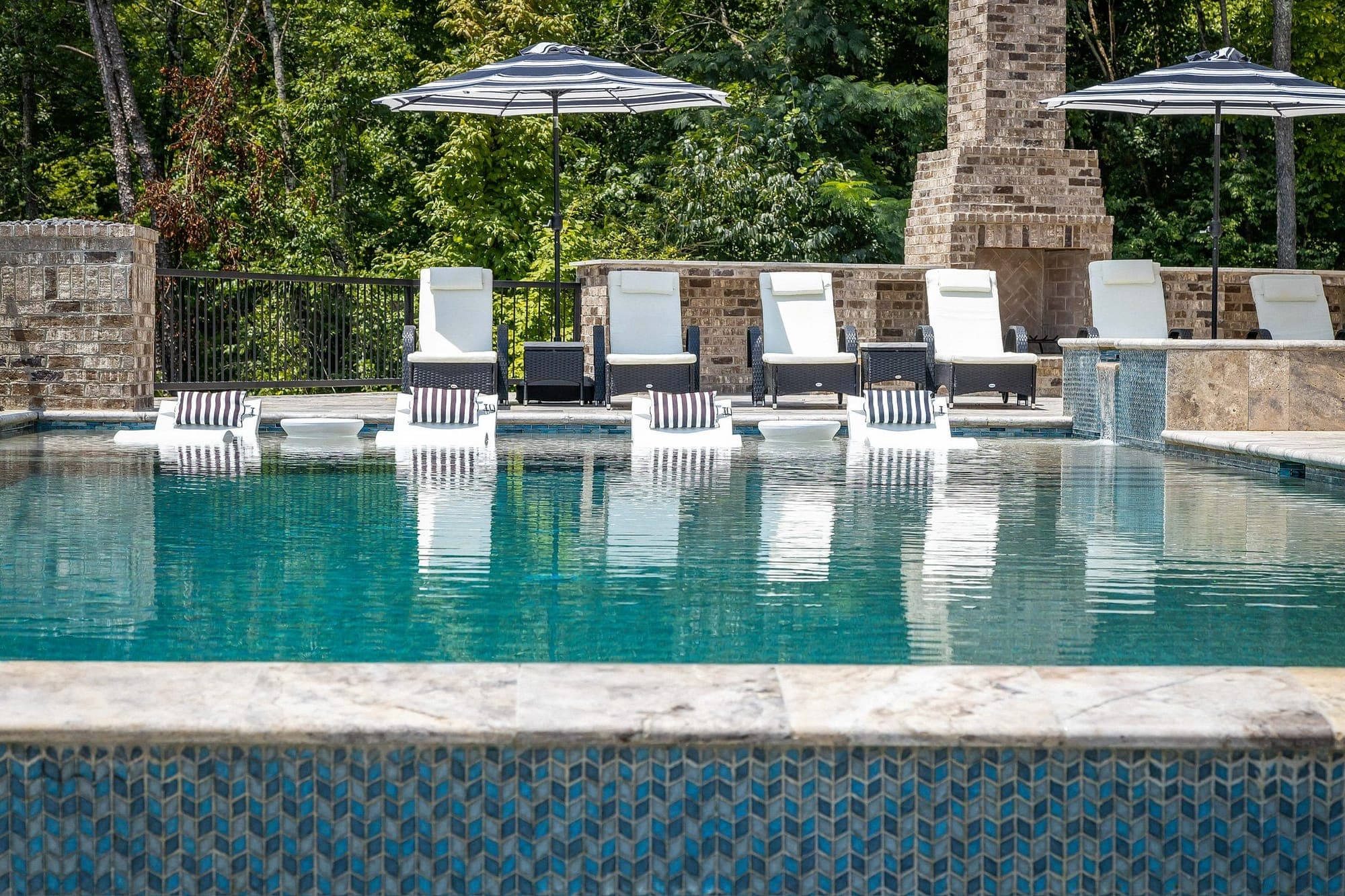 Pool with a tanning ledge of white loungers, striped umbrellas, a brick outdoor fireplace, and a blue mosaic tile wall