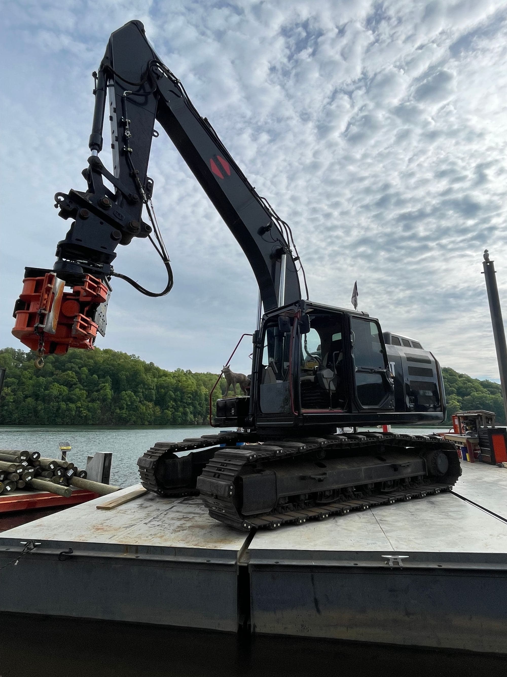 DOCK&DECK excavator with an orange vibratory pile-driving head on a steel work barge, with a stack of round wood pilings beside it on an East Tennessee lake